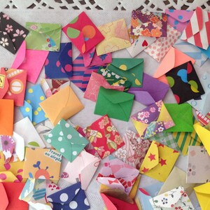 May include: A collection of colorful, patterned paper envelopes of various sizes. The envelopes are arranged in a random pattern on a white lace tablecloth.