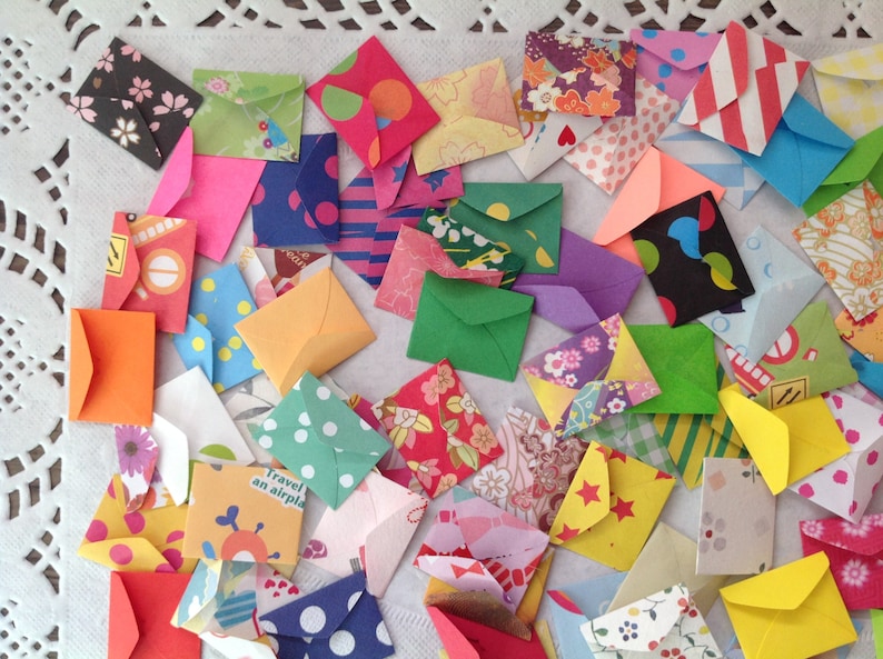 May include: A collection of colorful, patterned paper envelopes of various sizes. The envelopes are arranged in a random pattern on a white lace tablecloth.