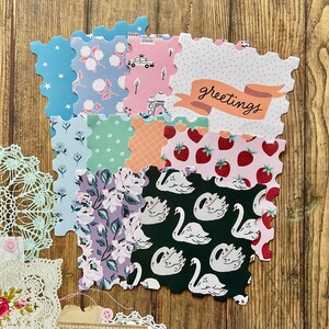 May include: A selection of decorative paper cutouts with scalloped edges, showcasing diverse patterns such as floral designs, strawberries, swans, and polka dots. One cutout displays the word "greetings". The cutouts are arranged on a wooden surface.