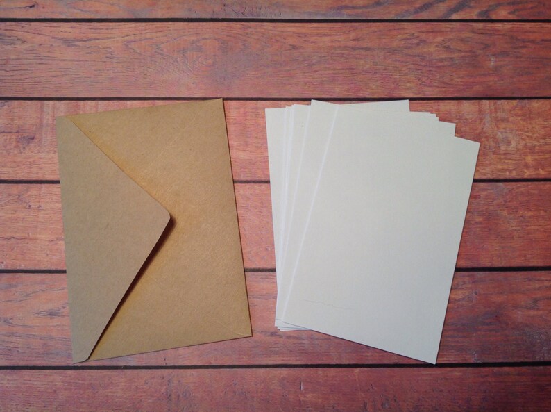 Peut inclure: Une enveloppe en papier brun et une pile de papier cartonn&eacute; blanc sur une surface en bois.