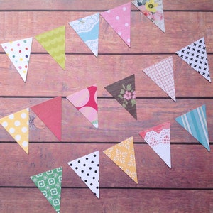 Può includere: Una ghirlanda di bandierine di carta a forma triangolare, di colori vivaci e con diversi motivi, tra cui pois, strisce, disegni floreali e forme geometriche. Le bandierine sono appese su uno sfondo di legno.