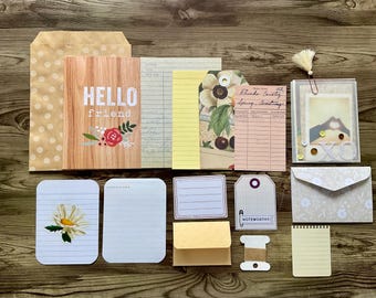 Op de afbeelding: Een verzameling papierknutselbenodigdheden, waaronder enveloppen, kaartjes en decoratief papier met een bloemenpatroon. De kaartjes hebben gestreept papier en één kaartje heeft de tekst "Hello Friend" erop gedrukt.