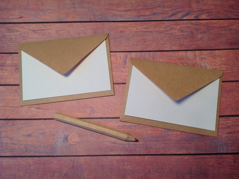 Peut inclure: Deux enveloppes brunes avec des int&eacute;rieurs blancs sont pos&eacute;es sur une surface en bois. Un crayon en bois est pos&eacute; &agrave; c&ocirc;t&eacute; des enveloppes.