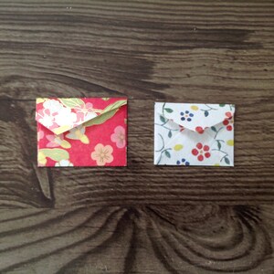 May include: Two small, folded envelopes with floral patterns. The envelope on the left has a red background with white and gold flowers. The envelope on the right has a white background with blue, yellow, and red flowers.