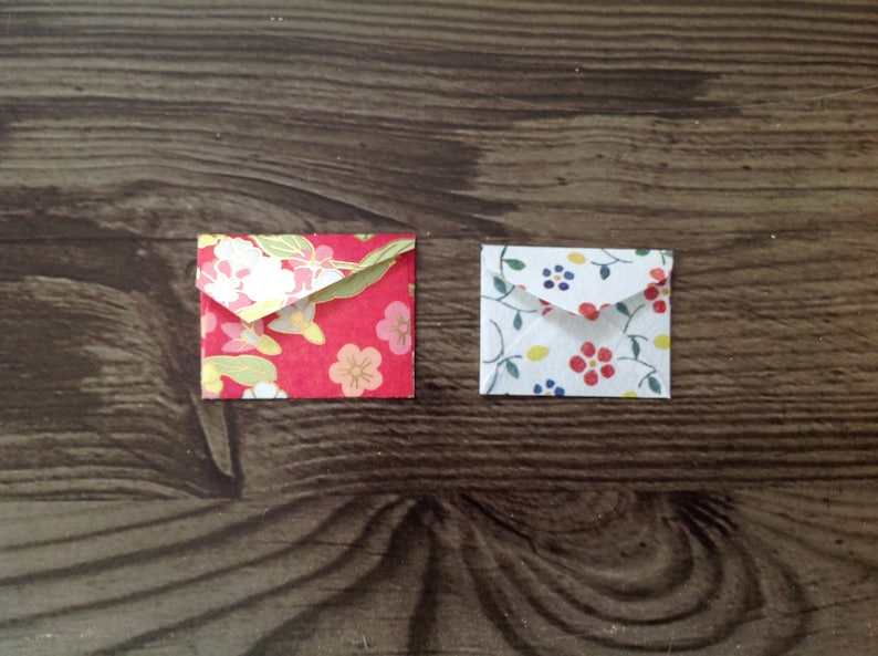 May include: Two small, folded envelopes with floral patterns. The envelope on the left has a red background with white and gold flowers. The envelope on the right has a white background with blue, yellow, and red flowers.