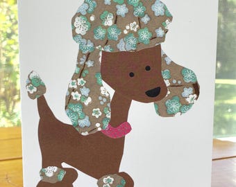 May include: A white greeting card featuring a brown poodle illustration. The dog has a brown body, a pink collar, and brown ears and paws decorated with a light blue and white floral pattern. The poodle also wears a matching floral hat.