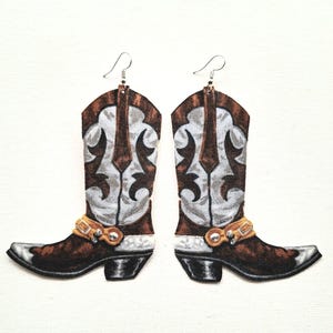 Könnte beinhalten: Ein Paar Cowboy-Stiefel-Ohrringe. Die Ohrringe sind braun, weiß und silberfarben mit einem dekorativen Design. Die Stiefel haben eine silberne Spitze und einen goldfarbenen Riemen um den Knöchel. Die Ohrringe hängen an silbernen Haken.