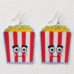 Puede incluir: Un par de pendientes de cubo de palomitas de maíz a rayas rojas y blancas con una cara de dibujos animados. El cubo de palomitas de maíz tiene una parte superior amarilla y una parte inferior azul. La cara tiene dos ojos negros, una ceja negra en cada ojo y una boca negra.