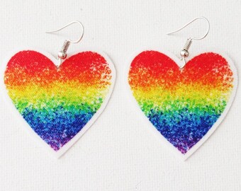 Könnte beinhalten: Ein Paar regenbogenförmige Ohrringe in Herzform. Die Ohrringe sind aus weißem Material mit einem Regenbogenmuster. Die Ohrringe hängen an silbernen Haken.