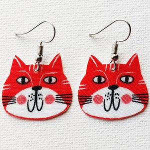 Puede incluir: Un par de pendientes de gato rojo con ganchos plateados. Las caras de los gatos tienen pelaje blanco, ojos negros y bigotes negros.