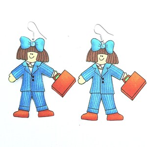 May include: A pair of novelty earrings featuring a cartoon businesswoman design. Each earring depicts a figure in a blue pinstripe suit, holding an orange briefcase, with a blue bow and brown hair. The earrings have silver-colored hooks.
