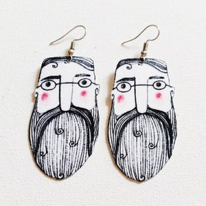 May include: A pair of dangle earrings featuring a cartoon illustration of a man with a beard, glasses, and pink cheeks. The earrings are made of fabric and have silver hooks.