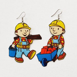 May include: A pair of novelty earrings featuring cartoon construction workers. Each earring depicts a worker in a yellow hard hat, blue overalls, and a red and yellow plaid shirt. One worker carries a wooden beam, the other pushes a blue wheelbarrow filled with red bricks.