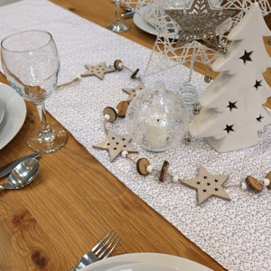 May include: A white wooden table with a white and grey patterned table runner. The table is set with two place settings, each with a white plate, a clear glass, and cutlery. A white ceramic Christmas tree with cut-out stars is on the table, along with a clear glass globe with a white candle inside. A string of wooden stars and beads is draped across the table runner.