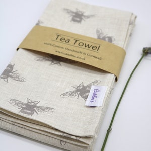 Pode incluir: Um conjunto de toalhas de ch&aacute; de linho bege com um padr&atilde;o de abelhas cinzas. As toalhas s&atilde;o dobradas e amarradas com uma faixa de papel castanho que diz "Tea Towel 100% Cotton. Handmade in Cornwall. www.caitlins.co.uk".