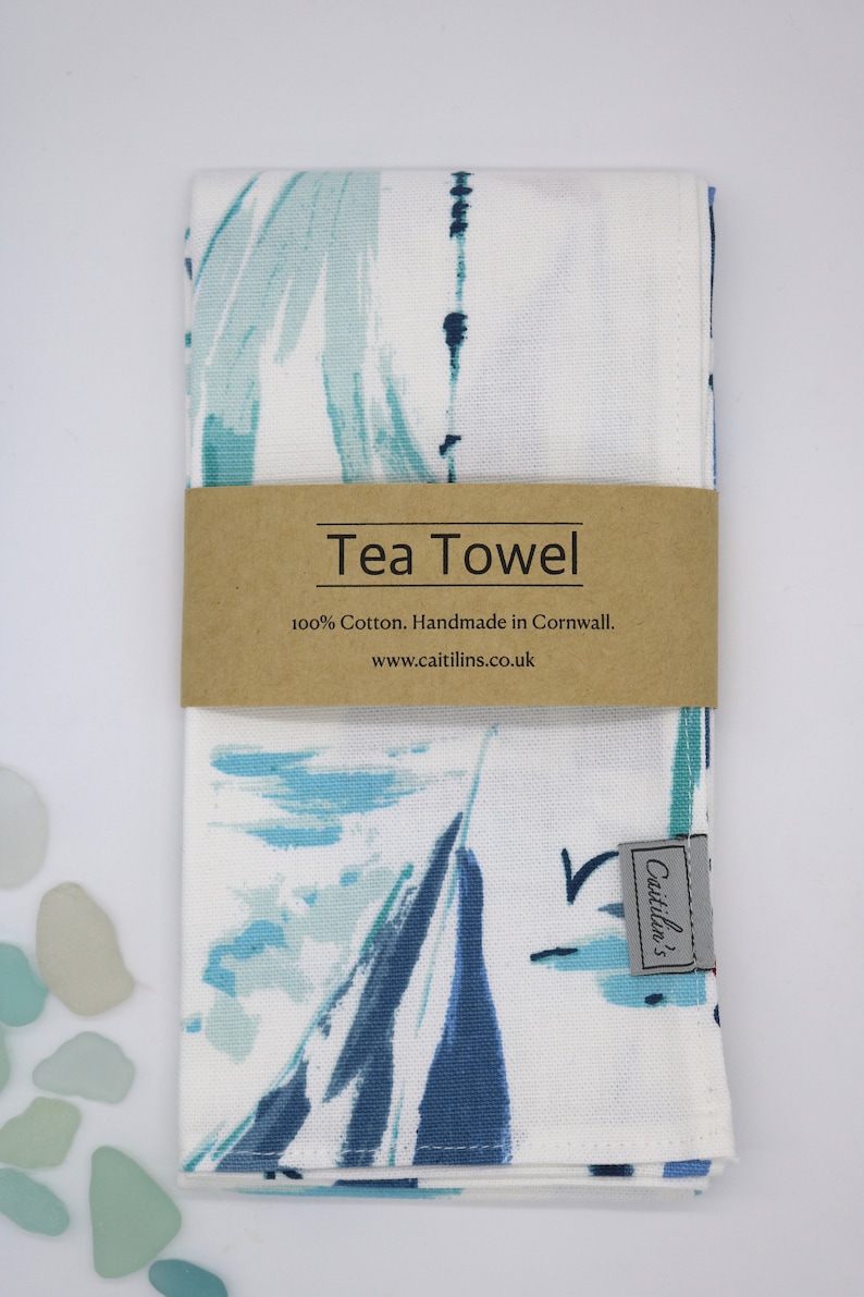 Pode incluir: Uma toalha de ch&aacute; branca com um design abstrato azul e verde. A toalha tem um r&oacute;tulo castanho que diz "Tea Towel 100% Cotton. Handmade in Cornwall. www.caitlins.co.uk" e um r&oacute;tulo cinzento mais pequeno que diz "Caitlin's".