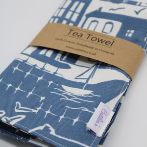 Pode incluir: Uma toalha de ch&aacute; azul e branca com um desenho de casas e barcos. A toalha est&aacute; dobrada e tem uma etiqueta marrom que diz "Tea Towel 100% Cotton. Handmade in Cornwall. www.caitlins.co.uk".