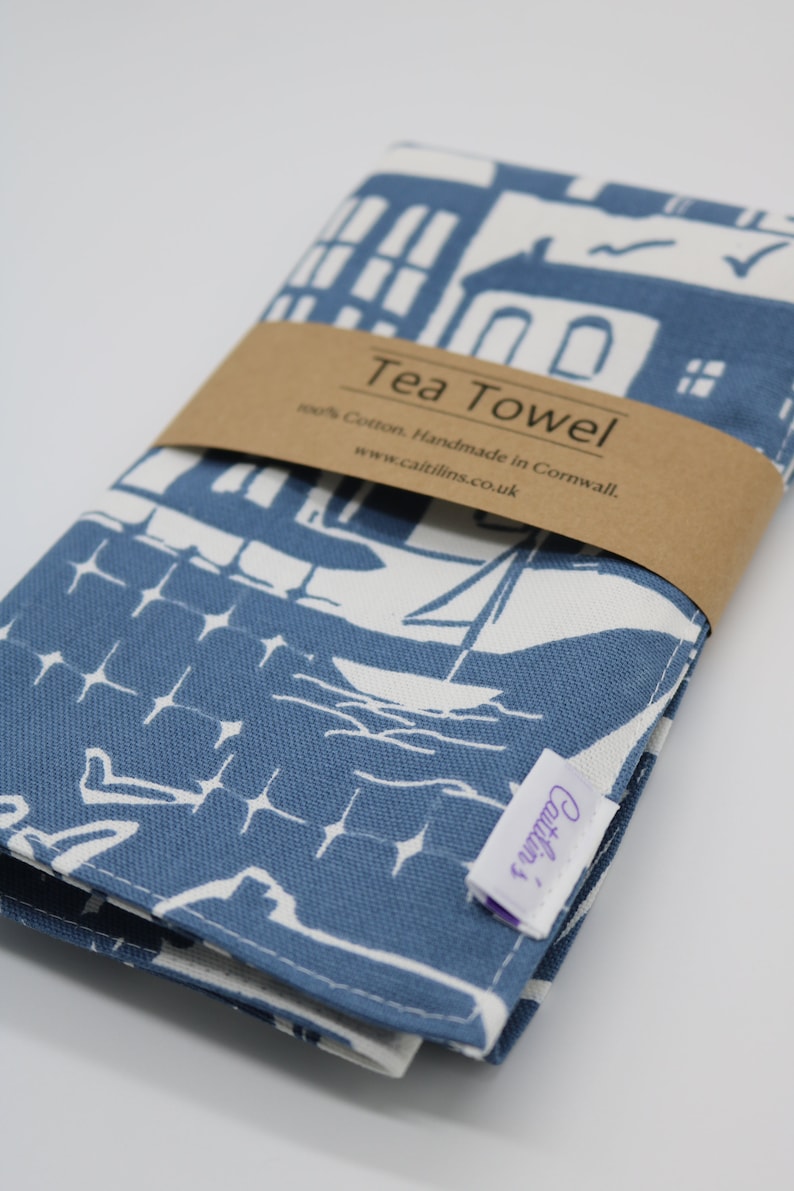 Pode incluir: Uma toalha de ch&aacute; azul e branca com um desenho de casas e barcos. A toalha est&aacute; dobrada e tem uma etiqueta marrom que diz "Tea Towel 100% Cotton. Handmade in Cornwall. www.caitlins.co.uk".