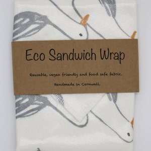 May include: A white fabric sandwich wrap with a gray and orange unicorn design. The wrap is folded and has a brown label that reads "Eco Sandwich Wrap" with the text "Reusable, vegan friendly and food safe fabric. Handmade in Cornwall."