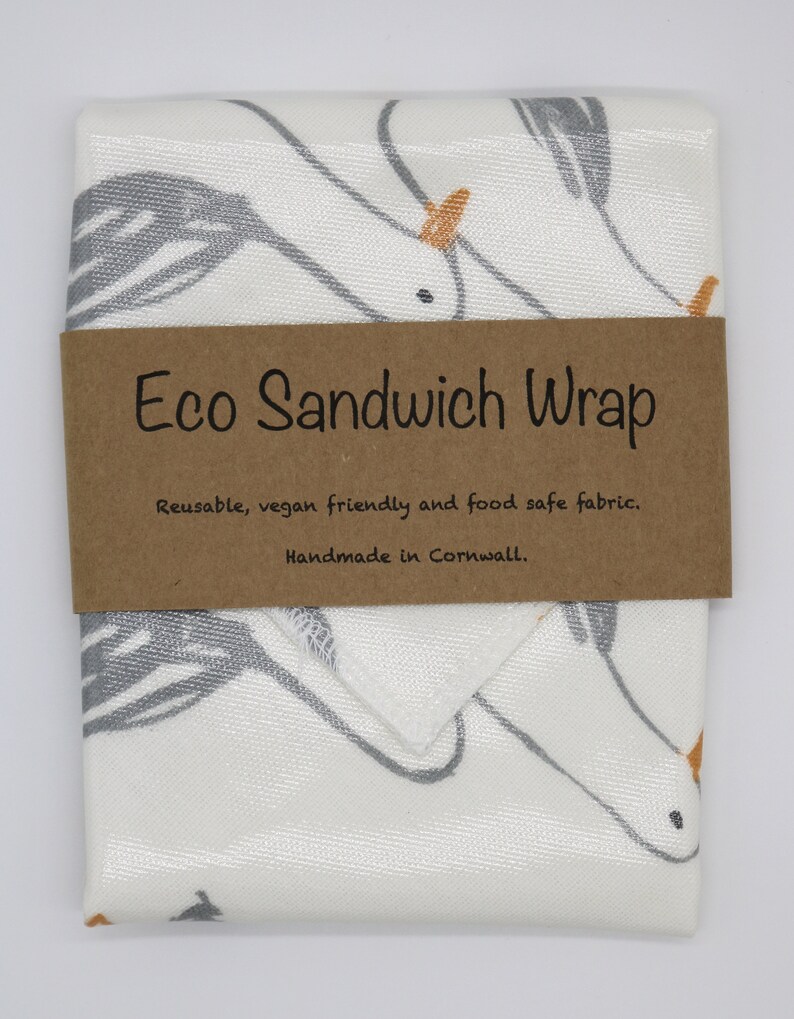 May include: A white fabric sandwich wrap with a gray and orange unicorn design. The wrap is folded and has a brown label that reads "Eco Sandwich Wrap" with the text "Reusable, vegan friendly and food safe fabric. Handmade in Cornwall."
