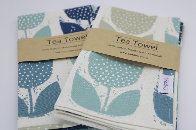 Pode incluir: Duas toalhas de ch&aacute; com um design floral. As toalhas est&atilde;o dobradas e empilhadas uma em cima da outra. A toalha superior &eacute; turquesa com bolinhas brancas e a toalha inferior &eacute; azul com bolinhas brancas. As toalhas s&atilde;o etiquetadas com "Tea Towel" e "100% Algod&atilde;o. Feito &agrave; m&atilde;o na Cornualha. www.caitilins.co.uk".