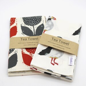 Pode incluir: Duas toalhas de ch&aacute;, uma com um design floral vermelho e cinza e a outra com um design de papagaio-do-mar branco e cinza. Ambas as toalhas t&ecirc;m o texto "Tea Towel" e "100% Algod&atilde;o. Feito &agrave; m&atilde;o na Cornualha. www.caitilins.co.uk" impresso nelas.