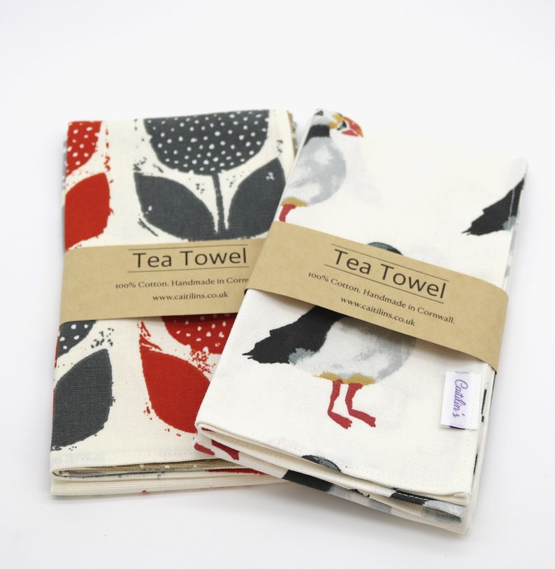 Pode incluir: Duas toalhas de ch&aacute;, uma com um design floral vermelho e cinza e a outra com um design de papagaio-do-mar branco e cinza. Ambas as toalhas t&ecirc;m o texto "Tea Towel" e "100% Algod&atilde;o. Feito &agrave; m&atilde;o na Cornualha. www.caitilins.co.uk" impresso nelas.