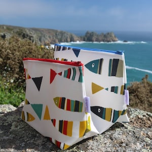May include: Three white zippered pouches with colorful fish designs. The pouches are lined up on a rocky surface with a blue ocean and sky in the background. The pouches have the text "Childer's" on them.