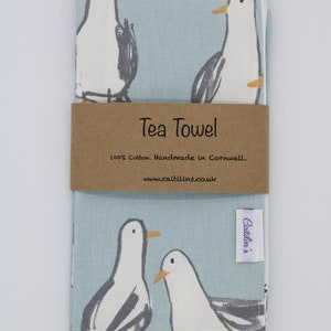 Pode incluir: Uma toalha de ch&aacute; azul claro com uma borda branca e um padr&atilde;o de gaivotas. A toalha tem um r&oacute;tulo castanho que diz "Tea Towel 100% Cotton. Handmade in Cornwall, www.caitlins.co.uk" e um r&oacute;tulo roxo que diz "Caitlin's".