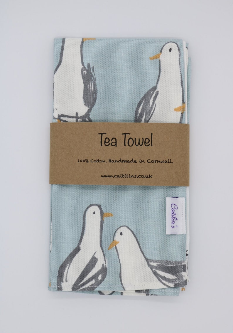 Pode incluir: Uma toalha de ch&aacute; azul claro com uma borda branca e um padr&atilde;o de gaivotas. A toalha tem um r&oacute;tulo castanho que diz "Tea Towel 100% Cotton. Handmade in Cornwall, www.caitlins.co.uk" e um r&oacute;tulo roxo que diz "Caitlin's".