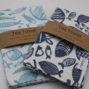 Pode incluir: Duas toalhas de ch&aacute; com fundo branco e um padr&atilde;o de peixes azuis. O texto na etiqueta marrom diz "Tea Towel 100% Cotton. Handmade in Cornwall. www.caitilins.co.uk".