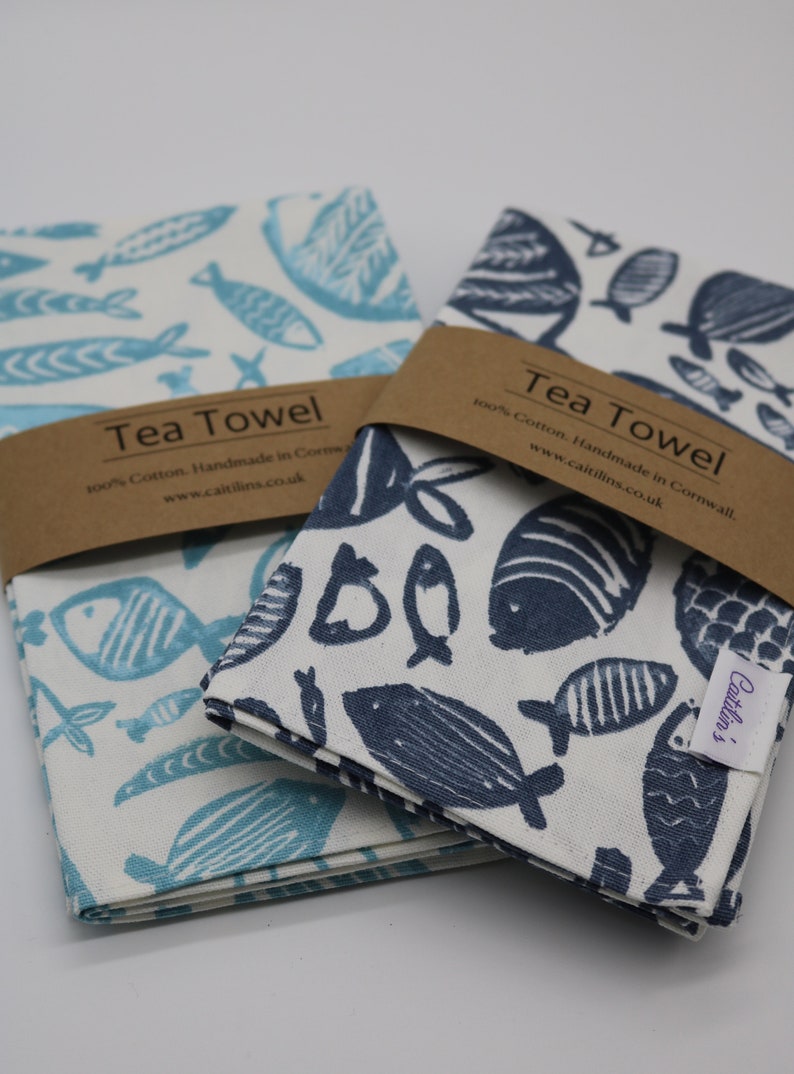 Pode incluir: Duas toalhas de ch&aacute; com fundo branco e um padr&atilde;o de peixes azuis. O texto na etiqueta marrom diz "Tea Towel 100% Cotton. Handmade in Cornwall. www.caitilins.co.uk".