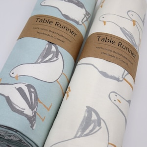 May include: Two rolled up table runners with a seagull pattern. The table runner on the left is light blue with white seagulls. The table runner on the right is white with grey seagulls. Both table runners have a brown label that says "Table Runner 100% cotton. Backed table runner. Handmade in Cornwall."