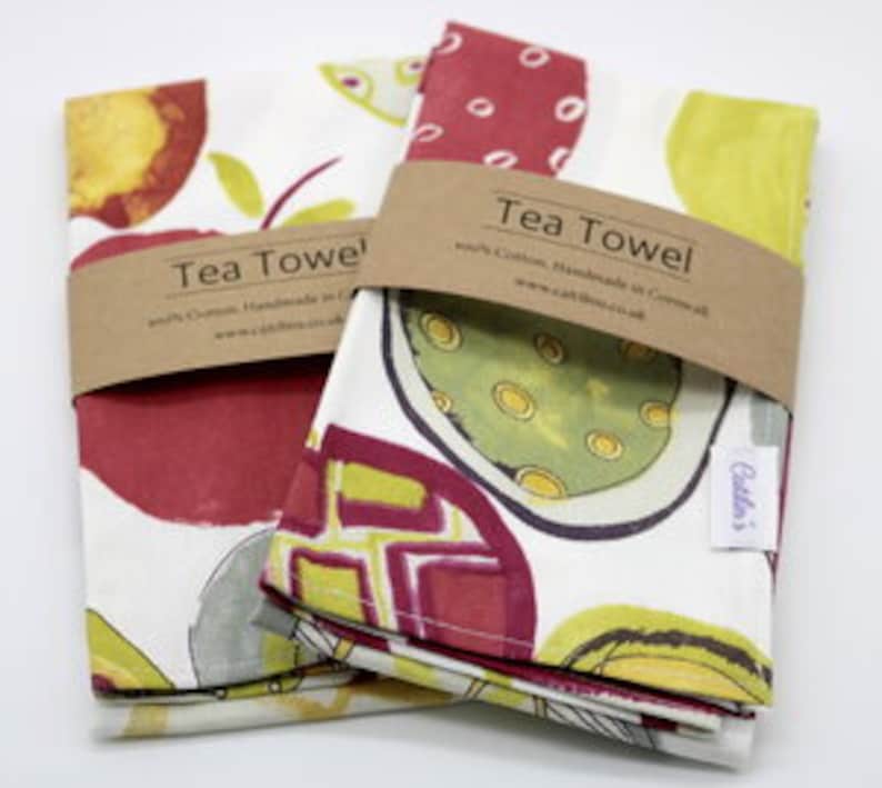 Pode incluir: Duas toalhas de ch&aacute; dobradas com um design abstrato de frutas coloridas. As toalhas s&atilde;o brancas com ilustra&ccedil;&otilde;es de frutas vermelhas, amarelas e verdes. As toalhas s&atilde;o etiquetadas com "Tea Towel" e o texto "100% Algod&atilde;o, Feito &agrave; m&atilde;o na Cornualha, www.cottonandco.co.uk".