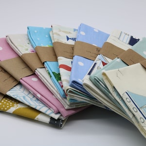 Pode incluir: Uma pilha de toalhas de ch&aacute; com v&aacute;rios padr&otilde;es e cores, incluindo azul, rosa e amarelo. As toalhas est&atilde;o dobradas e embrulhadas em papel castanho com o texto "Tea Towels" impresso nelas.