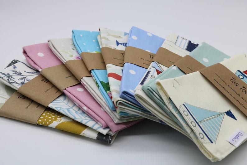 Pode incluir: Uma pilha de toalhas de ch&aacute; com v&aacute;rios padr&otilde;es e cores, incluindo azul, rosa e amarelo. As toalhas est&atilde;o dobradas e embrulhadas em papel castanho com o texto "Tea Towels" impresso nelas.