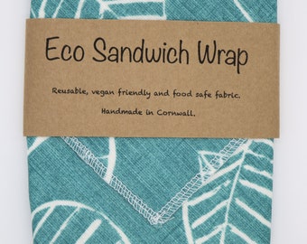May include: A teal blue fabric sandwich wrap with a white leaf pattern. The wrap has a brown paper label that reads "Eco Sandwich Wrap" and "Reusable, vegan friendly and food safe fabric. Handmade in Cornwall."