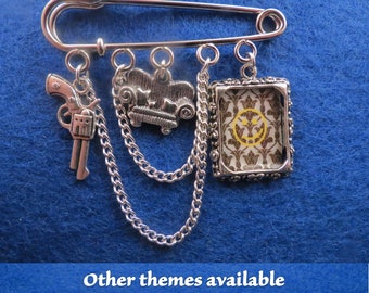May include: A silver safety pin with multiple charms attached, including a gun, a couch, and a framed image with a yellow smiley face on a brown and gold patterned background.  The charms are connected to the pin with silver chains.