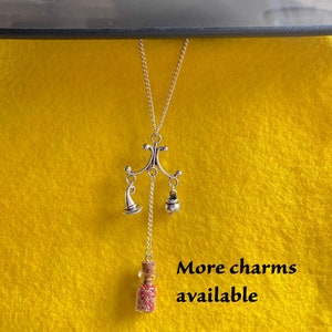 May include: Silver necklace with a witch hat, cauldron, and a small glass bottle charm filled with red glitter. The text "More charms available" is written below the necklace.