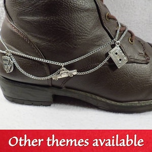 Puede incluir: Botas de cuero marrón con cadenas y dijes plateados. Los dijes incluyen un coche, una cinta de casete y un logotipo.