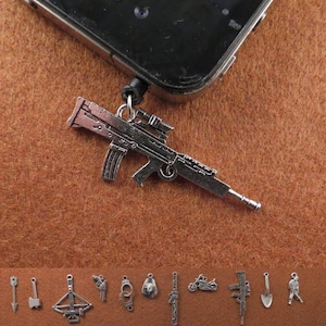 Puede incluir: Un amuleto plateado de un rifle con mira telescópica, unido a una funda de teléfono negra.