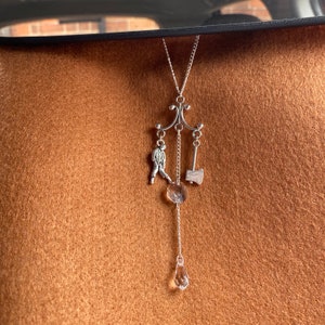 May include: A silver car charm with a dangling figure, an axe, and a crystal pendant. The charm is hanging from a silver chain.