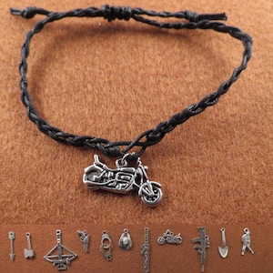 Op de afbeelding: Zwart gevlochten koordarmband met een zilveren motorfietsbedel. De armband ligt op een bruine ondergrond.