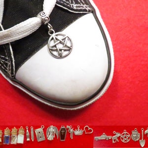 May include: A silver pentagram charm attached to a white shoelace on a black and white trainer. The charm is hanging from a silver bead. The shoe is on a red surface. Below the shoe are various silver charms, including a pair of angel wings, a bottle with a cork, a bottle with blue liquid, a bottle with a cork, a bottle with white liquid, a sword, a cassette tape, an angel, a vampire, a coffin with a cross, a crown, a heart, a car, a moose, a pentagram, a shield, a shovel, a squirrel, and a tie.