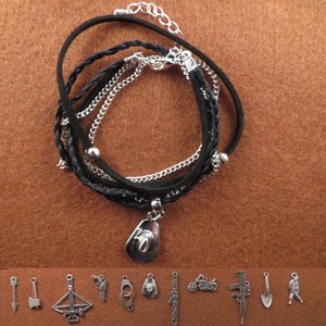 Può includere: Un braccialetto in pelle nera con più strati di cordoncino intrecciato e catena d'argento. Il braccialetto ha un ciondolo d'argento a forma di cappello da cowboy appeso ad esso. L'immagine mostra anche una varietà di altri ciondoli d'argento, tra cui una freccia, un'ascia, una balestra, una pistola, manette, un cappello, un coltello, una motocicletta, un fucile, una pala e una persona.