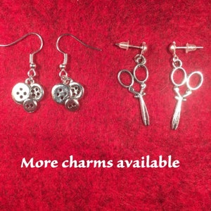 May include: Three pairs of silver earrings. The first pair features a charm of three stacked buttons. The second pair features a charm of a pair of scissors. The third pair also features a charm of a pair of scissors. The text "More charms available" is at the bottom of the image.