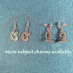 May include: Two pairs of silver earrings. One pair features a paint palette charm, the other pair features a microscope charm. The text "More subject charms available" is at the bottom of the image.