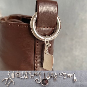 Puede incluir: Un bolso de mano de cuero marrón con un dije plateado sujeto al asa. El dije tiene forma de cuchillo y tiene una cadena plateada unida a él.