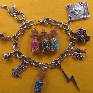 May include: A silver charm bracelet with various charms, including a snake, a book, a spider, a lightning bolt, and small bottles filled with colorful beads. The bracelet is on a yellow background.