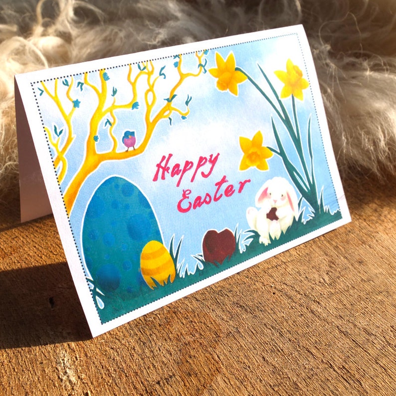 May include: A white card with a blue sky background and a yellow daffodil flower. The card says "Happy Easter" in pink letters. There is a white bunny holding a chocolate egg and a blue egg with yellow accents.